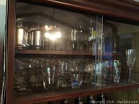 Glassware and Coca Cola collectible bottles displayed on shelves inside a bookcase behind glass doors.
