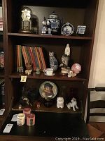 Full shelf showing books, animal figurines, candles and decorative plates, framed portrait plate, and clock.