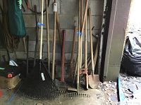 Wide angle photo showing various yard tools leaning against a wall including rakes, axes, hoes, a hole digger, and a small shovel.