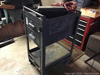 Metal rolling cart with two drawers, bottom shelf, and items on top in a basement setting.
