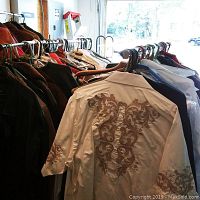 Two racks of men's vintage jackets, suits, coats and shirts shown hanging on metal racks. Clothing is mixed colors with embroidery and patterns visible.
