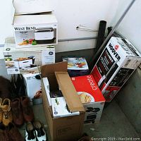 Photo showing boxed kitchen appliances stacked together including West Bend slow cooker box, GE deep fryer box, Hoover Floormax vacuum cleaner box, GE 4-in-1 grill, Ninja cooking system, Dansk Ultimate Pot, and Morrone turkey fryer box.