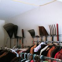 Wide shot showing variety of yard tools hanging on wall rack including leaf rakes, shovels, pitchfork, and hedge trimmers above a clothing rack.