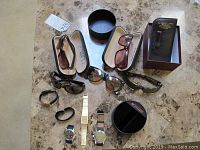 Photo showing 5 men's watches and 4 pairs of sunglasses arranged on a marble surface: dark framed sunglasses, brown lens sunglasses in cases, Seiko watch without band, Movado stainless steel watch, Austin gold-tone watch, and other watches.