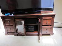 Front view of wooden entertainment center with TV on top, center glass-door cabinet open to show AV equipment inside, drawers on the sides