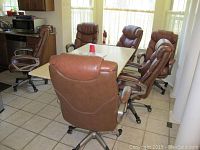 Six brown leather-like upholstered office chairs arranged around a table in a kitchen area. Chairs have padded armrests and caster wheels.