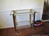 Full view of metal and glass bar cart with visible wear, showing structure, two glass shelves, casters, placed on wood floor against wall.