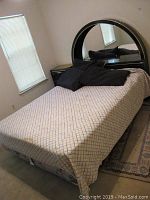 Full view of bed showing headboard with mirror, mattress with patterned bedspread, black pillows, and metal bed frame visible beneath mattress.