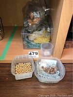 Photo of 3 plastic containers on a shelf containing assorted beads in gold, silver, and light brown colors along with a small clear cup nearby.