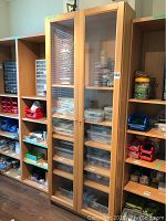Tall laminate cabinet with two glass front doors showing internal shelving and plastic storage containers inside. Cabinet has a light wood finish and metal handles.