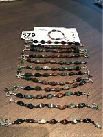 Twelve multi-colored oval-beaded bracelets laid out on a flat surface, showing various bead colors and toggle clasps with charms.