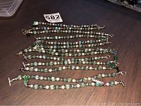 Full view of over a dozen bracelets laid out containing textured green, blue, and white beads with toggle clasps and ribbon charms.