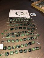 View of all 10 bracelets laid flat with Barbara brand paper and lot tag visible, showing uniform green and black stone square design with toggle clasps.