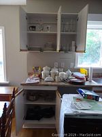 Kitchen view with open shelves, showing ceramic canister set, dinner and bread plates, mugs, pots and fry pans, butter dish, electric kettle and boxes on countertop.