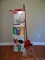 Photo showing white bookshelf with humidifier box on top, broom, cleaning supplies on shelves, small iron on floor
