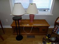 Photo of solid wood coffee table with single door and two pink ceramic base table lamps with white pleated shades, placed on the coffee table and floor beside it, in front of a window.