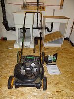 Full view of mower on plywood floor with manual, oil bottles and gas cans behind