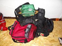 Stacked collection of various bags including black backpacks, green travel bag, and red duffel bag on a plywood floor