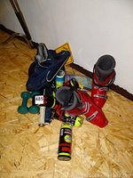Photo showing paired red ski boots, tennis rackets, cans of tennis balls, green dumbbells, blue ball hopper bag, and yellow caution sign on floor.