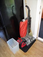 Older model Kirby upright vacuum with red cloth bag, wooden broom behind, compact red and black handheld vacuum on floor next to a light gray plastic trash can, black door mat beneath them.