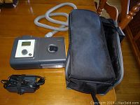 Full lot displayed on wooden table: CPAP machine with hose attached, charger coiled, and black carrying bag beside.