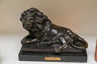 Frontal view of bronze lion seated on marble base with plaque reading “LION.”
