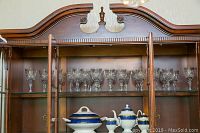 Group of about 10 crystal wine glasses with cut pattern inside a wood display cabinet on upper shelves.