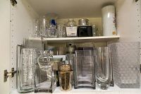 Two shelves mounted in a white cabinet filled with assorted glass and ceramic vases in multiple shapes and finishes including clear, frosted, silver, black, and white.