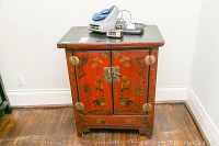 Front view of red Asian style vintage cabinet showing painted figures, floral motifs, brass hardware, and latch, and some objects placed on top