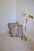 Photo showing the 46 inch gold-tone floor lamp with adjustable head placed next to two square decorative pillows with striped fabric and ruffled edges against a white wall and carpeted floor.