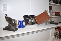 Photo showing metal elephant figurine, blue ceramic mug, pottery piece, corkscrew set in leather case, and candle holder on mantle