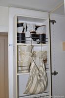 Full view of white shelving unit containing six black lamp shades with gold trim on top shelf and six white ceramic scalloped planter pots below, striped fabric bag hanging in front.
