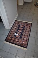 The rectangular mat laid flat on a tiled floor showing front side with geometric and floral patterns in navy, beige, and rust colors with tasseled edges.