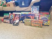 Wide view of assortment showing boxed games and puzzles on carpet in front of sofa. Includes Cranium, Cadoo, Battleship, Clue, Monopoly, various puzzles, and smaller games like Checkers and 5 Second Game.