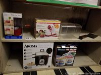 Photo shows shelf with six kitchen items including boxed Aroma rice cooker, Donvier ice cream maker, Starfrit apple peeler, measuring sifter, clear bread box, and a large cleaver.