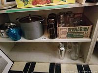 Shelf shelf with large Circulon metal stock pot with lid, metal serving bowls, Boston shaker, glass martini pitcher, and box of Bernardin mason jars