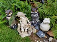 Full shot of garden decor set showing girl figurine with two cats, two owl statues, white fat cat figurine, and natural garden stones among ferns.