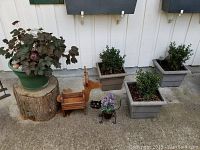 View of wooden box planters with small shrubs, green plastic planter with plant, wood reindeer, metal cat plant holder on a sidewalk against a white wooden wall.