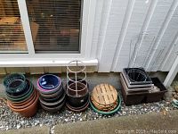 Assorted plant pots stacked outside along the house wall, including terracotta and plastic pots, some with metal wire cages and plant supports.