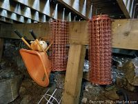 Photo shows orange wheelbarrow with rake and shovel inside, red garden fence rolls hanging, and two orange tomato cages hanging on wood beam.