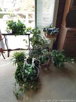 Wide view of assortment of live and faux plants along with metal plant stand and decorative cat sculpture, arranged on carpet near window.