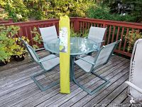 Full patio set showing round glass-top table with four chairs and upright green umbrella bag at center on wooden deck.
