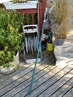 Outdoor photo of garden hose coiled on a white plastic reel, and a long green pole pruner standing upright with a rope for operation.