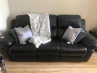 Front view of the black leather Cenro reclining couch with decorative pillows and a throw blanket draped on it, placed against a white wall on a wood floor.