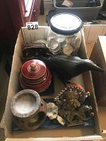 Photo showing lot contents in cardboard box including black cast iron crow side view, red and gold lidded urn, blue plate, jar with wax tea lights, oriental brass figurines, minerals, and coasters.