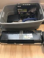 Plastic bin containing Asus RT-N12 Wireless-N300 router box, Linksys router box, power cables, and other components with a black keyboard or pad box in front.