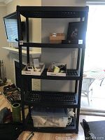 Full view of black plastic shelving unit with four shelves, placed indoors near sliding glass door and fireplace.