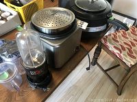 Photo of Cuisinart rice cooker, Magic Bullet blender, and T-fal fryer on wooden table with chairs in background