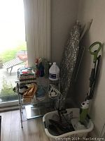 Photo showing metal shelving unit containing cleaning supplies, woven baskets, iron, cords, and analog scale, next to laundry basket and ironing board near a window.