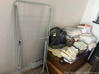 Photo showing white metal drying rack leaning against wall next to a wooden chest holding the Bissell SpotClean Complete spot cleaner and a stack of folded towels and linens.